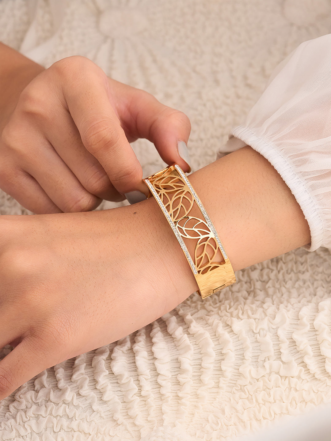 Person wearing a gold bracelet with leaf design on a textured beige surface